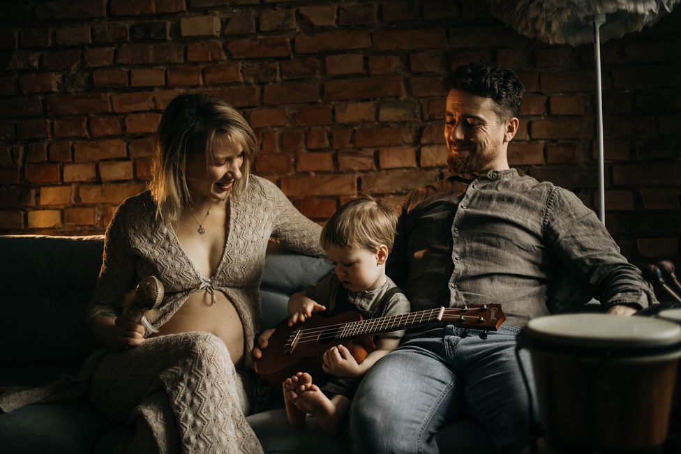 Sesja ciążowa Ostrów Wielkopolski - kobieta z brzuszkiem i jej mąż siedzą na kapapie wraz z małym synkiem