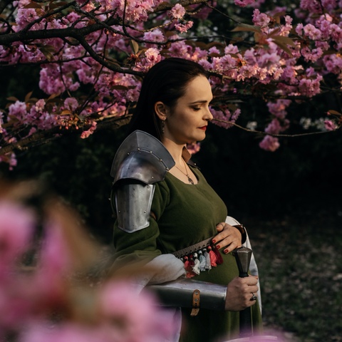 Sesja ciążowa - kobieta w historycznym stroju z rękoma złożonymi na brzuchu pośród kwiecących drzew