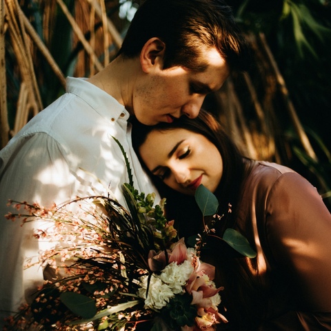 Sesja zdjęciowa - para przytula się do siebie pod rozświetlonym lasem wraz z bukietem kwiatów