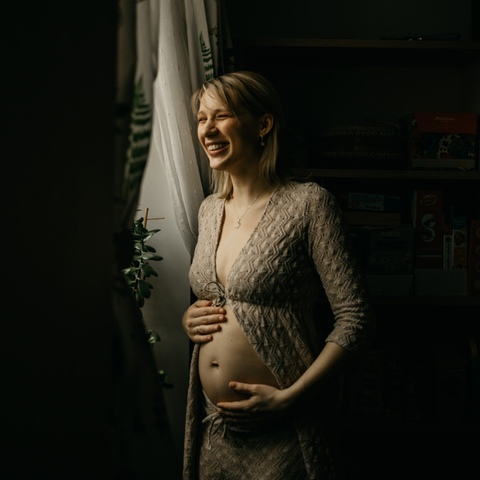 Sesja ciążowa - uśmiechnięta kobieta stojąca przy oknie